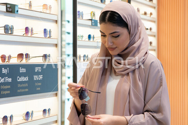 تسوق وشراء اكسسوارات نسائية عصرية أنيقة، متابعة آخر صيحات الموضة والجمال، متعة التسوق في مولات الإمارات، امرأة سعودية تقوم بمعاينة النظارات الملونة، شابة عربية خليجية اماراتية محجبة ترتدي عباءة تقوم باختيار نظارة شمسية ملائمة لملامح الوجه