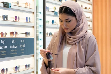 تسوق وشراء اكسسوارات نسائية عصرية أنيقة، متابعة آخر صيحات الموضة والجمال، متعة التسوق في مولات الإمارات، امرأة سعودية تقوم بمعاينة النظارات الملونة، شابة عربية خليجية اماراتية محجبة ترتدي عباءة تقوم باختيار نظارة شمسية ملائمة لملامح الوجه