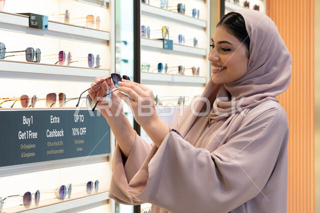 السعادة والمتعة بالتسوق في مولات الإمارات، مواكبة آخر صيحات الموضة والجمال، أحدث الاكسسوارات النسائية العصرية الأنيقة، صورة مقربة لشابة عربية خليجية اماراتية محجبة ترتدي عباءة تقوم باختيار نظارة شمسية ملائمة لملامح الوجه،  امرأة سعودية تقوم بمعاينة النظارات