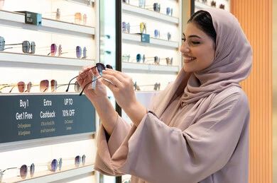 السعادة والمتعة بالتسوق في مولات الإمارات، مواكبة آخر صيحات الموضة والجمال، أحدث الاكسسوارات النسائية العصرية الأنيقة، صورة مقربة لشابة عربية خليجية اماراتية محجبة ترتدي عباءة تقوم باختيار نظارة شمسية ملائمة لملامح الوجه،  امرأة سعودية تقوم بمعاينة النظارات