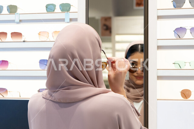 اكسسوارات نسائية عصرية، اختيار نظارة شمسيه مناسبة، التأثير الإيجابي للتسوق على المرأة، الاهتمام بالمظهر الخارجي، امرأة اماراتية تقوم بتجربة النظارات قبل الشراء، صورة مقربة من الخلف لشابة عربية خليجية سعودية محجبة ترتدي عباءة تقوم بقياس نظاره ورؤيتها على المرآة