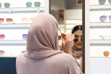 اكسسوارات نسائية عصرية، اختيار نظارة شمسيه مناسبة، التأثير الإيجابي للتسوق على المرأة، الاهتمام بالمظهر الخارجي، امرأة اماراتية تقوم بتجربة النظارات قبل الشراء، صورة مقربة من الخلف لشابة عربية خليجية سعودية محجبة ترتدي عباءة تقوم بقياس نظاره ورؤيتها على المرآة