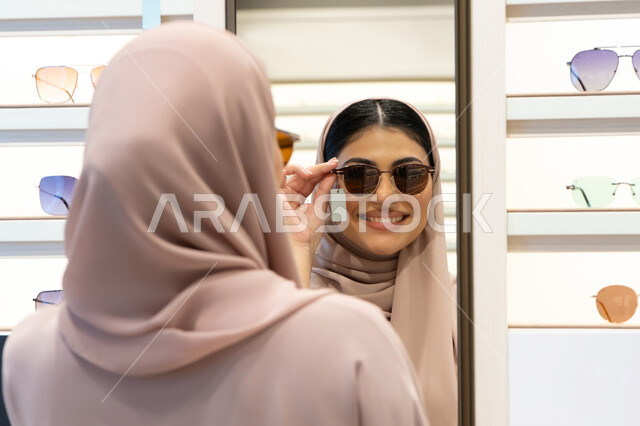متعة التسوق في الإمارات، اكسسوارات نسائية عصرية، اختيار نظارة شمسيه مناسبة، الاهتمام بالمظهر الخارجي، امرأة اماراتية تقوم بتجربة النظارات قبل الشراء، صورة مقربة من الخلف لشابة عربية خليجية سعودية محجبة ترتدي عباءة تقوم بقياس نظاره ورؤيتها على المرآة