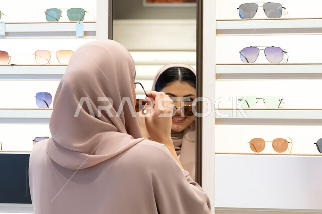 النظرإلى المرآة بتعابير السعادة والسرور، الاستمتاع بالتسوق في الإمارات،  اكسسوارات نسائية عصرية، اختيار نظارة شمسيه مناسبة، امرأة اماراتية تقوم بتجربة النظارات قبل الشراء، صورة مقربة من الخلف لشابة عربية خليجية سعودية محجبة ترتدي عباءة تقوم بقياس نظاره