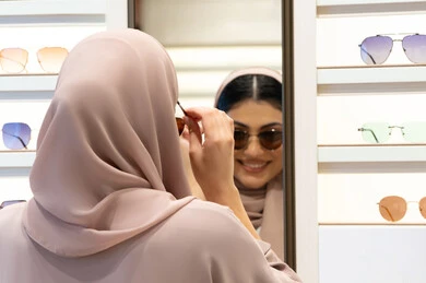 Expressions of happiness and pleasure, enjoying shopping in the Emirates, fashionable women's accessories, choosing suitable sunglasses, an Emirati woman trying on glasses before buying, a close-up from the back of a veiled Saudi Arabian Gulf young woman wearing an abaya trying on her glasses and looking at them in the mirror
