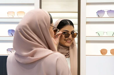 اختيار نظارة شمسيه مناسبة، التأثير الإيجابي للتسوق على المرأة، اكسسوارات نسائية عصرية، الاهتمام بالمظهر الخارجي، امرأة اماراتية تقوم بتجربة النظارات قبل الشراء، صورة مقربة من الخلف لشابة عربية خليجية سعودية محجبة ترتدي عباءة تقوم بقياس نظاره ورؤيتها على المرآة
