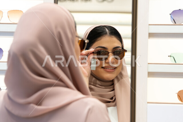 الاستمتاع بالتسوق في الإمارات، الاهتمام بالمظهر الخارجي،  اكسسوارات نسائية عصرية، اختيار نظارة شمسيه مناسبة، امرأة اماراتية تقوم بتجربة النظارات قبل الشراء، صورة مقربة من الخلف لشابة عربية خليجية سعودية محجبة ترتدي عباءة تقوم بقياس نظاره ورؤيتها على المرآة