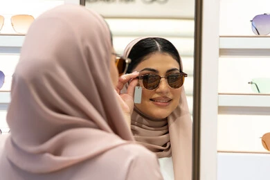 الاستمتاع بالتسوق في الإمارات، الاهتمام بالمظهر الخارجي،  اكسسوارات نسائية عصرية، اختيار نظارة شمسيه مناسبة، امرأة اماراتية تقوم بتجربة النظارات قبل الشراء، صورة مقربة من الخلف لشابة عربية خليجية سعودية محجبة ترتدي عباءة تقوم بقياس نظاره ورؤيتها على المرآة