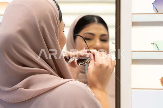 اختيار نظارة شمسيه مناسبة، الاستمتاع بالتسوق في الإمارات،  اكسسوارات نسائية عصرية، امرأة اماراتية تقوم بتجربة النظارات قبل الشراء، صورة مقربة من الخلف لشابة عربية خليجية سعودية محجبة ترتدي عباءة تقوم بقياس نظاره