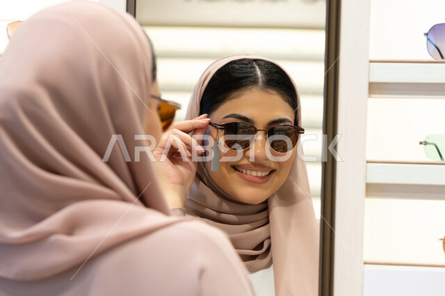 متعة التسوق في الإمارات، اكسسوارات نسائية عصرية، اختيار نظارة شمسيه مناسبة، الاهتمام بالمظهر الخارجي، امرأة اماراتية تقوم بتجربة النظارات قبل الشراء، صورة مقربة من الخلف لشابة عربية خليجية سعودية محجبة ترتدي عباءة تقوم بقياس نظاره ورؤيتها على المرآة