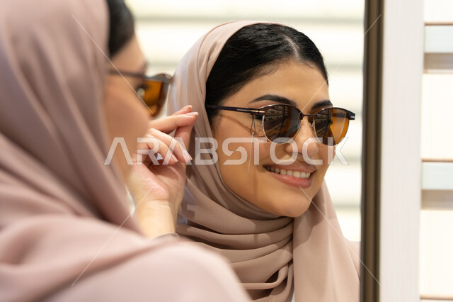 اكسسوارات نسائية عصرية، اختيار نظارة شمسيه مناسبة، التأثير الإيجابي للتسوق على المرأة، الاهتمام بالمظهر الخارجي، امرأة اماراتية تقوم بتجربة النظارات قبل الشراء، صورة مقربة من الخلف لشابة عربية خليجية سعودية محجبة ترتدي عباءة تقوم بقياس نظاره ورؤيتها على المرآة