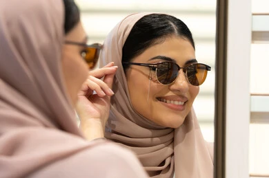اكسسوارات نسائية عصرية، اختيار نظارة شمسيه مناسبة، التأثير الإيجابي للتسوق على المرأة، الاهتمام بالمظهر الخارجي، امرأة اماراتية تقوم بتجربة النظارات قبل الشراء، صورة مقربة من الخلف لشابة عربية خليجية سعودية محجبة ترتدي عباءة تقوم بقياس نظاره ورؤيتها على المرآة