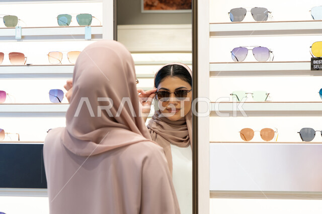 الاهتمام بالمظهر الخارجي، متعة التسوق في الإمارات، اكسسوارات نسائية عصرية، اختيار نظارة شمسيه مناسبة، امرأة اماراتية تقوم بتجربة النظارات قبل الشراء، صورة مقربة من الخلف لشابة عربية خليجية سعودية محجبة ترتدي عباءة تقوم بقياس نظاره ورؤيتها على المرآة