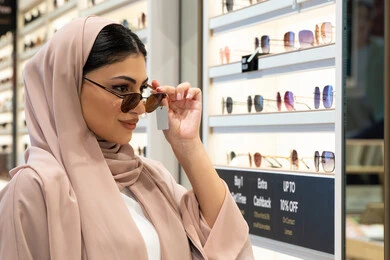 أرفف عرض النظارات داخل المحل، اختيار نظارة شمسيه مناسبة، الاستمتاع بالتسوق في الإمارات،  اكسسوارات نسائية عصرية، امرأة اماراتية تقوم بتجربة النظارات قبل الشراء، صورة مقربة لشابة عربية خليجية سعودية محجبة ترتدي عباءة تقوم بقياس نظاره