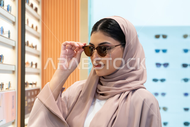 اقتناء واختيار نظارة شمسيه مناسبة، تشكيلة اكسسوارات نسائية عصرية أنيقة، الاستمتاع بالتسوق في الإمارات، امرأة اماراتية تقوم بتجربة النظارات قبل الشراء، صورة مقربة لشابة عربية خليجية سعودية محجبة ترتدي عباءة تقوم بقياس نظاره