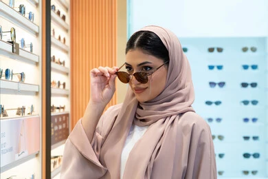 أرفف عرض النظارات داخل المحل، اختيار نظارة شمسيه مناسبة، الاستمتاع بالتسوق في الإمارات،  اكسسوارات نسائية عصرية، امرأة اماراتية تقوم بتجربة النظارات قبل الشراء، صورة مقربة لشابة عربية خليجية سعودية محجبة ترتدي عباءة تقوم بقياس نظاره