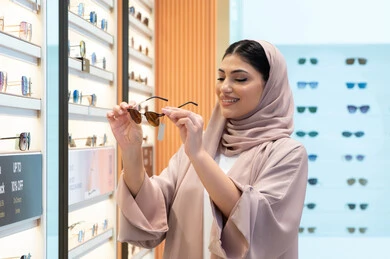 السعادة والمتعة بالتسوق في مولات الإمارات، مواكبة آخر صيحات الموضة والجمال، أحدث الاكسسوارات النسائية العصرية الأنيقة، صورة مقربة لشابة عربية خليجية اماراتية محجبة ترتدي عباءة تقوم باختيار نظارة شمسية ملائمة لملامح الوجه،  امرأة سعودية تقوم بمعاينة النظارات