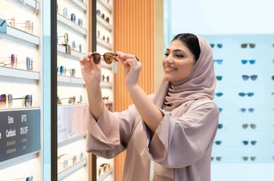 مواكبة آخر صيحات الموضة والجمال، الاهتمام بالمظهر الخارجي، أحدث الاكسسوارات النسائية، المتعة بالتسوق في مولات الإمارات، شابة عربية خليجية اماراتية محجبة ترتدي عباءة تقوم باختيار نظارة شمسية ملائمة لملامح الوجه،  امرأة سعودية تقوم بمعاينة النظارات