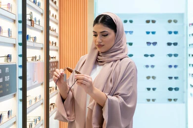 مواكبة آخر صيحات الموضة والجمال، الاهتمام بالمظهر الخارجي، أحدث الاكسسوارات النسائية، المتعة بالتسوق في مولات الإمارات، شابة عربية خليجية اماراتية محجبة ترتدي عباءة تقوم باختيار نظارة شمسية ملائمة لملامح الوجه،  امرأة سعودية تقوم بمعاينة النظارات