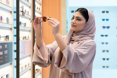 Emirati store selling the latest women's accessories, enjoy shopping in the UAE malls, keep up with the latest fashion and beauty trends, a close-up side view of a veiled Emirati Gulf Arab young woman wearing an abaya choosing sunglasses that suit her facial features, a Saudi woman examining the glasses