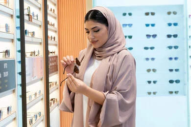 شراء اكسسوارات نسائية عصرية أنيقة، متابعة آخر صيحات الموضة والجمال، متعة التسوق في مولات الإمارات، امرأة سعودية تقوم بمعاينة النظارات الملونة، شابة عربية خليجية اماراتية محجبة ترتدي عباءة تقوم باختيار نظارة شمسية ملائمة لملامح الوجه