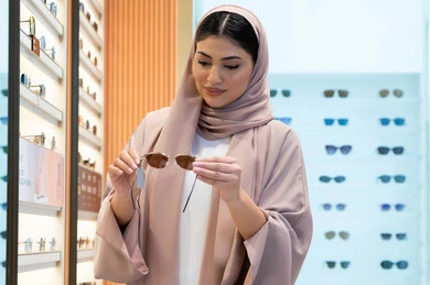 تسوق وشراء اكسسوارات نسائية عصرية أنيقة، متابعة آخر صيحات الموضة والجمال، متعة التسوق في مولات الإمارات، امرأة سعودية تقوم بمعاينة النظارات الملونة، شابة عربية خليجية اماراتية محجبة ترتدي عباءة تقوم باختيار نظارة شمسية ملائمة لملامح الوجه