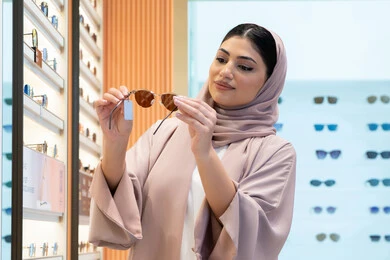 متابعة آخر صيحات الموضة والجمال، متعة التسوق في مولات الإمارات، اكسسوارات نسائية عصرية أنيقة، صورة مقربة لامرأة سعودية تقوم بمعاينة النظارات الملونة، شابة عربية خليجية اماراتية محجبة ترتدي عباءة تقوم باختيار نظارة شمسية ملائمة لملامح الوجه
