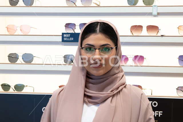 تعابير الثقة بالنفس، مفهوم ضعف النظر،الشراء من المحلات الإماراتية، اختيار نظارة ملائمة لملامح الوجه،  اقتناء نظارات طبية جديدة، صورة مقربة لشابة عربية خليجية سعودية محجبة ترتدي عباءة تنظر إلى الكاميرا بإيماءات السعادة، امرأة إماراتية تقوم بمعاينة النظارات