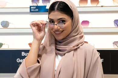 الشراء من المحلات الإماراتية، اختيار نظارة ملائمة لملامح الوجه، مفهوم ضعف النظر، اقتناء نظارات طبية جديدة، صورة مقربة لشابة عربية خليجية سعودية محجبة ترتدي عباءة تنظر إلى الكاميرا بإيماءات السعادة، امرأة إماراتية تقوم بمعاينة النظارات