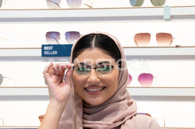 اختيار نظارة ملائمة لملامح الوجه، مفهوم ضعف النظر، النظر إلى الكاميرا بإيماءات السعادة، اقتناء نظارات طبية جديدة، صورة مقربة لشابة عربية خليجية سعودية محجبة ترتدي عباءة تقوم بوضع النظارة قبل الشراء، امرأة إماراتية تقوم بمعاينة النظارات