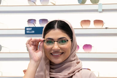 اختيار نظارة ملائمة لملامح الوجه، مفهوم ضعف النظر، النظر إلى الكاميرا بإيماءات السعادة، اقتناء نظارات طبية جديدة، صورة مقربة لشابة عربية خليجية سعودية محجبة ترتدي عباءة تقوم بوضع النظارة قبل الشراء، امرأة إماراتية تقوم بمعاينة النظارات