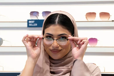 مفهوم ضعف النظر،الشراء من المحلات الإماراتية، اختيار نظارة ملائمة لملامح الوجه،  اقتناء نظارات طبية جديدة، صورة مقربة لشابة عربية خليجية سعودية محجبة ترتدي عباءة تنظر إلى الكاميرا بإيماءات السعادة، امرأة إماراتية تقوم بمعاينة النظارات