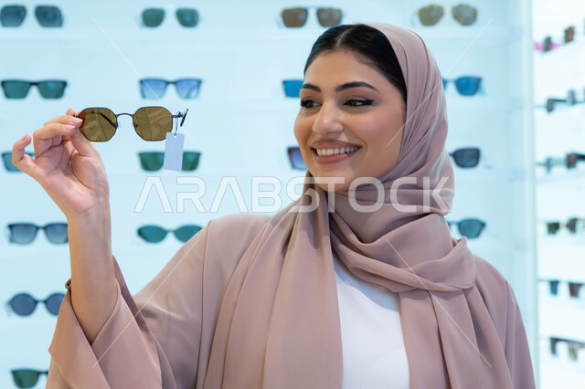السعادة والمتعة بالتسوق في مولات الإمارات، مواكبة آخر صيحات الموضة والجمال، أحدث الاكسسوارات النسائية العصرية الأنيقة، صورة مقربة لشابة عربية خليجية اماراتية محجبة ترتدي عباءة تقوم باختيار نظارة شمسية ملائمة لملامح الوجه،  امرأة سعودية تقوم بمعاينة النظارات