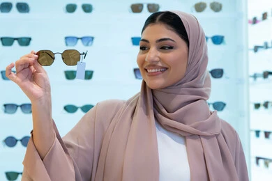 السعادة والمتعة بالتسوق في مولات الإمارات، مواكبة آخر صيحات الموضة والجمال، أحدث الاكسسوارات النسائية العصرية الأنيقة، صورة مقربة لشابة عربية خليجية اماراتية محجبة ترتدي عباءة تقوم باختيار نظارة شمسية ملائمة لملامح الوجه،  امرأة سعودية تقوم بمعاينة النظارات