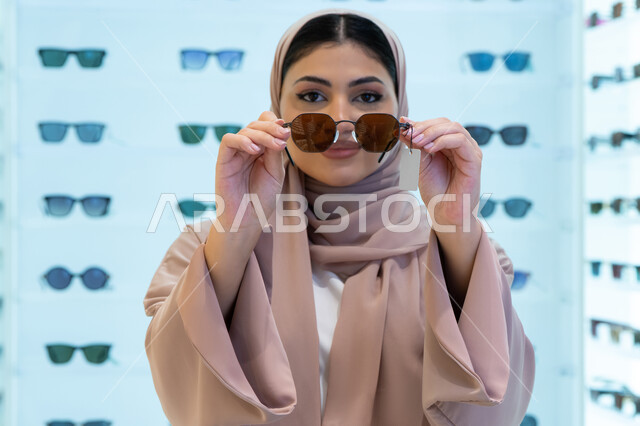 اقتناء واختيار نظارة شمسيه مناسبة، تشكيلة اكسسوارات نسائية عصرية أنيقة، الاستمتاع بالتسوق في الإمارات، امرأة اماراتية تقوم بتجربة النظارات قبل الشراء، صورة مقربة لشابة عربية خليجية سعودية محجبة ترتدي عباءة تقوم بقياس نظاره