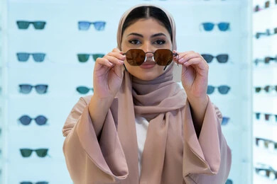 اقتناء واختيار نظارة شمسيه مناسبة، تشكيلة اكسسوارات نسائية عصرية أنيقة، الاستمتاع بالتسوق في الإمارات، امرأة اماراتية تقوم بتجربة النظارات قبل الشراء، صورة مقربة لشابة عربية خليجية سعودية محجبة ترتدي عباءة تقوم بقياس نظاره