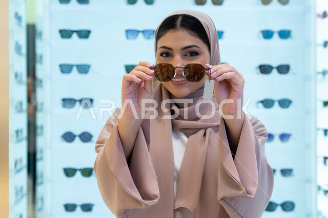 تشكيلة اكسسوارات نسائية عصرية أنيقة، أرفف عرض النظارات داخل المحل، اختيار نظارة شمسيه مناسبة، الاستمتاع بالتسوق في الإمارات، امرأة اماراتية تقوم بتجربة النظارات قبل الشراء، صورة مقربة لشابة عربية خليجية سعودية محجبة ترتدي عباءة تقوم بقياس نظاره