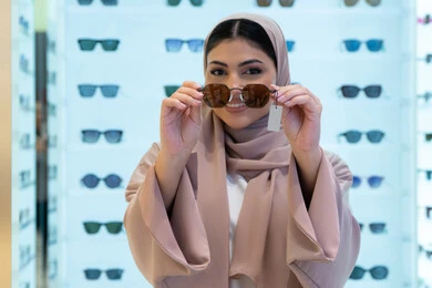 تشكيلة اكسسوارات نسائية عصرية أنيقة، أرفف عرض النظارات داخل المحل، اختيار نظارة شمسيه مناسبة، الاستمتاع بالتسوق في الإمارات، امرأة اماراتية تقوم بتجربة النظارات قبل الشراء، صورة مقربة لشابة عربية خليجية سعودية محجبة ترتدي عباءة تقوم بقياس نظاره
