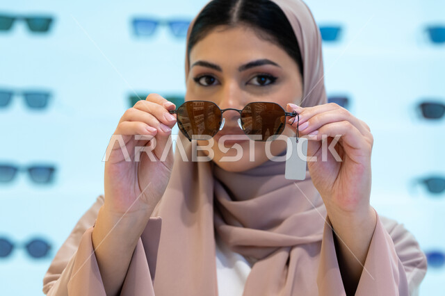 النظر إلى الكاميرا بإيماءات الثقة بالنفس، اختيار نظارة شمسية ملائمة لملامح الوجه، اكسسوارات نسائية عصرية أنيقة، الاستمتاع بالتسوق في الإمارات، امرأة اماراتية تقوم بتجربة النظارات قبل الشراء، صورة مقربة لشابة عربية خليجية سعودية محجبة ترتدي عباءة تقوم بوضع نظاره