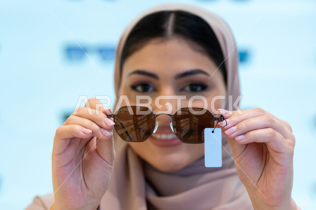 اكسسوارات نسائية عصرية أنيقة، النظر إلى الكاميرا بإيماءات السرور، اقتناء واختيار نظارة شمسيه مناسبة، الاستمتاع بالتسوق في الإمارات، امرأة اماراتية تقوم بتجربة النظارات قبل الشراء، صورة مقربة لشابة عربية خليجية سعودية محجبة ترتدي عباءة تقوم بقياس نظاره