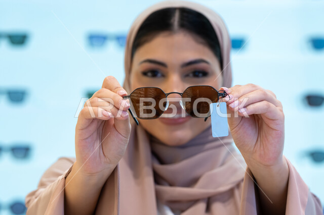 النظر إلى الكاميرا بإيماءات السرور، اقتناء واختيار نظارة شمسيه مناسبة، تشكيلة اكسسوارات نسائية عصرية أنيقة، الاستمتاع بالتسوق في الإمارات، امرأة اماراتية تقوم بتجربة النظارات قبل الشراء، صورة مقربة لشابة عربية خليجية سعودية محجبة ترتدي عباءة تقوم بقياس نظاره