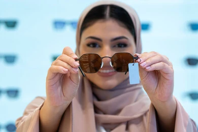 النظر إلى الكاميرا بإيماءات السرور، اقتناء واختيار نظارة شمسيه مناسبة، تشكيلة اكسسوارات نسائية عصرية أنيقة، الاستمتاع بالتسوق في الإمارات، امرأة اماراتية تقوم بتجربة النظارات قبل الشراء، صورة مقربة لشابة عربية خليجية سعودية محجبة ترتدي عباءة تقوم بقياس نظاره