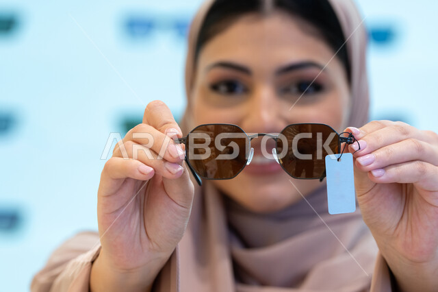 اختيار نظارة شمسية ملائمة لملامح الوجه، اكسسوارات نسائية عصرية أنيقة، النظر إلى الكاميرا بإيماءات السرور، الاستمتاع بالتسوق في الإمارات، امرأة اماراتية تقوم بتجربة النظارات قبل الشراء، صورة مقربة لشابة عربية خليجية سعودية محجبة ترتدي عباءة تقوم بقياس نظاره