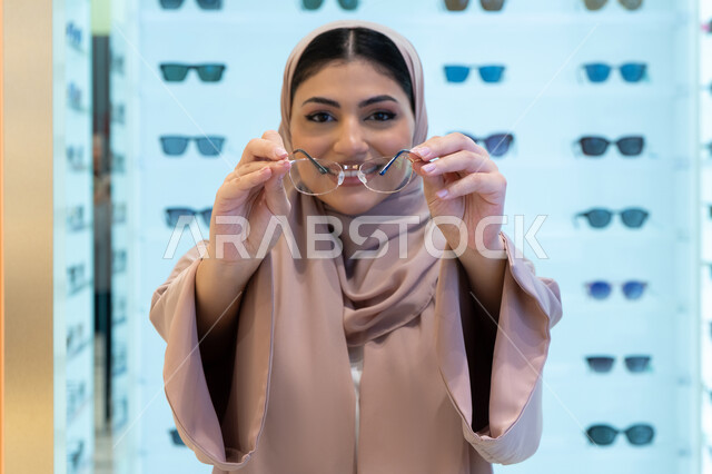التسوق في إحدى مولات الإمارات، تصحيح وتحسين الرؤية، مفهوم ضعف النظر، النظر إلى الكاميرا بإيماءات السعادة، اقتناء نظارات طبية جديدة، صورة مقربة لشابة عربية خليجية سعودية ترتدي عباءة تقوم باختيار نظارة ملائمة لملامح وجهها، امرأة إماراتية تقوم بمعاينة النظارات