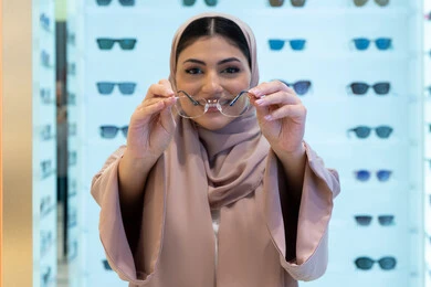 التسوق في إحدى مولات الإمارات، تصحيح وتحسين الرؤية، مفهوم ضعف النظر، النظر إلى الكاميرا بإيماءات السعادة، اقتناء نظارات طبية جديدة، صورة مقربة لشابة عربية خليجية سعودية ترتدي عباءة تقوم باختيار نظارة ملائمة لملامح وجهها، امرأة إماراتية تقوم بمعاينة النظارات