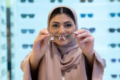 Looking at the camera with happy gestures, buying new eyeglasses, shopping in a mall in the Emirates, the concept of poor eyesight, a close-up of a veiled Saudi Arabian Gulf young woman wearing an abaya choosing eyeglasses that suit her facial features, an Emirati woman examining eyeglasses