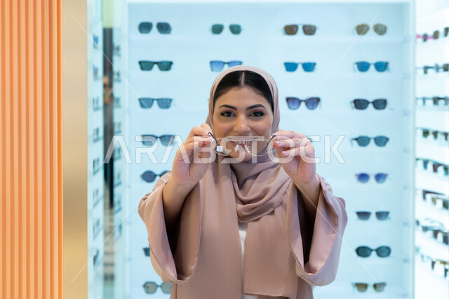 Correcting and improving vision, the concept of weak vision, looking at the camera with happy gestures, buying new eyeglasses, shopping in a mall in the Emirates, a close-up of a young Saudi Arabian Gulf woman wearing an abaya choosing eyeglasses that suit her facial features, an Emirati woman examining eyeglasses