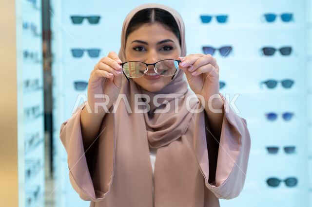 مفهوم ضعف النظر، النظر إلى الكاميرا بإيماءات السعادة، اقتناء نظارات طبية جديدة، التسوق في إحدى مولات الإمارات، صورة مقربة لشابة عربية خليجية سعودية محجبة ترتدي عباءة تقوم باختيار نظارة ملائمة لملامح وجهها، امرأة إماراتية تقوم بمعاينة النظارات