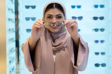 The concept of poor vision, looking at the camera with happy gestures, buying new eyeglasses, shopping in a mall in the Emirates, a close-up of a veiled Saudi Arabian Gulf young woman wearing an abaya choosing eyeglasses that suit her facial features, an Emirati woman examining eyeglasses