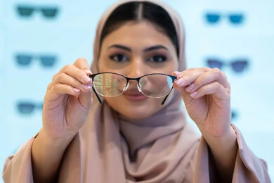 Buying new eyeglasses, shopping in a mall in the Emirates, the concept of poor eyesight, display shelves of products and accessories, a close-up of a veiled Saudi Arabian Gulf young woman wearing an abaya choosing eyeglasses that suit her facial features, an Emirati woman examining eyeglasses